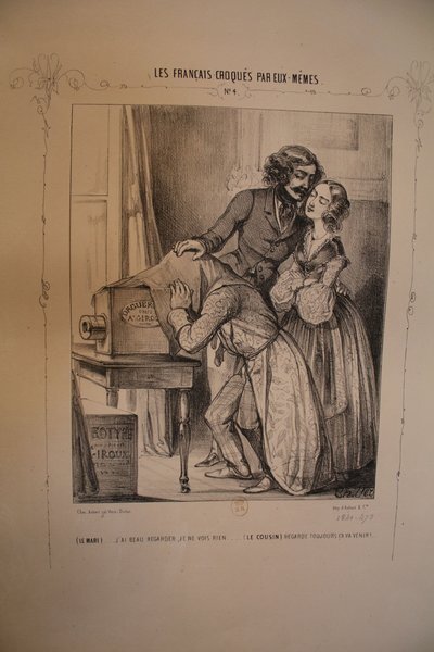 An illustration showing two women and a man in a 19th-century indoor setting. The man and one of the women are examining a large box that is labeled "Daguerreotype" resting on a small table. All individuals are dressed in detailed period clothing, suggestive of the July Monarchy era. The scene potentially relates to themes revolving around art and romance.