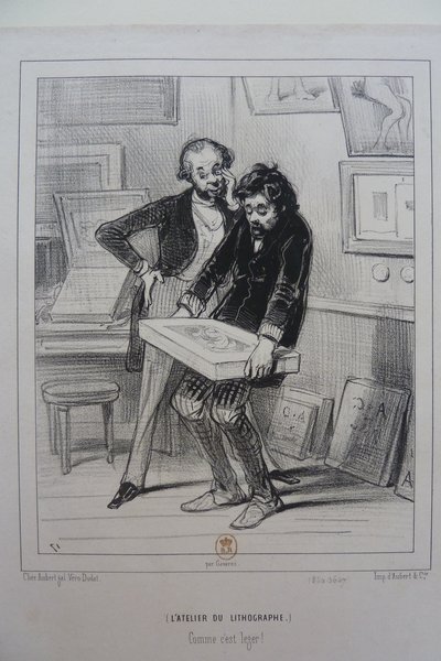 Two men in a studio, one holding a lithographic stone and the other with a monocle, near lithographic stones and prints during the July Monarchy. One man showcases facial hair and both appear as art critics or printmakers engaged in examining or discussing artwork in what seems to be a printmaking environment.