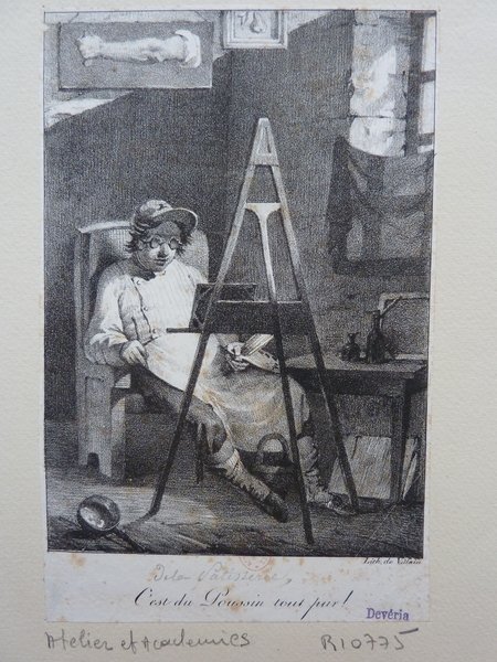 An artist wearing pince-nez sits by an easel in a studio, focusing on a painting. Surrounding the artist are various items including palettes, artists’ brushes, and a folio. There is a painting of an arm hung prominently above the artist.
